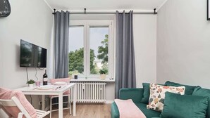 Ein Zimmer mit Esstisch, einem grünen Sofa, einem Fernseher und einem Fenster mit Vorhängen.