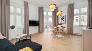 Una sala de estar moderna con un sofá, una mesa de comedor con sillas, un televisor y ventanas grandes con cortinas.
