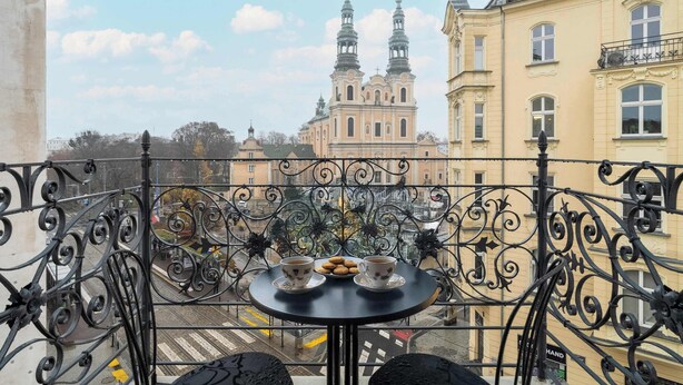 Un balcón con barandillas de hierro forjado ornamentadas, una mesa redonda con dos tazas y un plato de galletas, y vistas de un edificio histórico con dos torres.