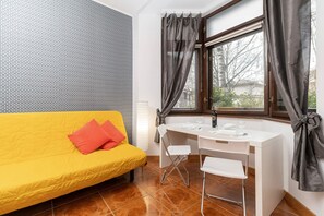 A room with a yellow sofa, a white desk with chairs, a window with curtains, and a patterned wall.