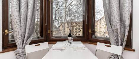 A dining table set for two with a bottle of wine, glasses, and plates, positioned in a room with large windows and curtains.