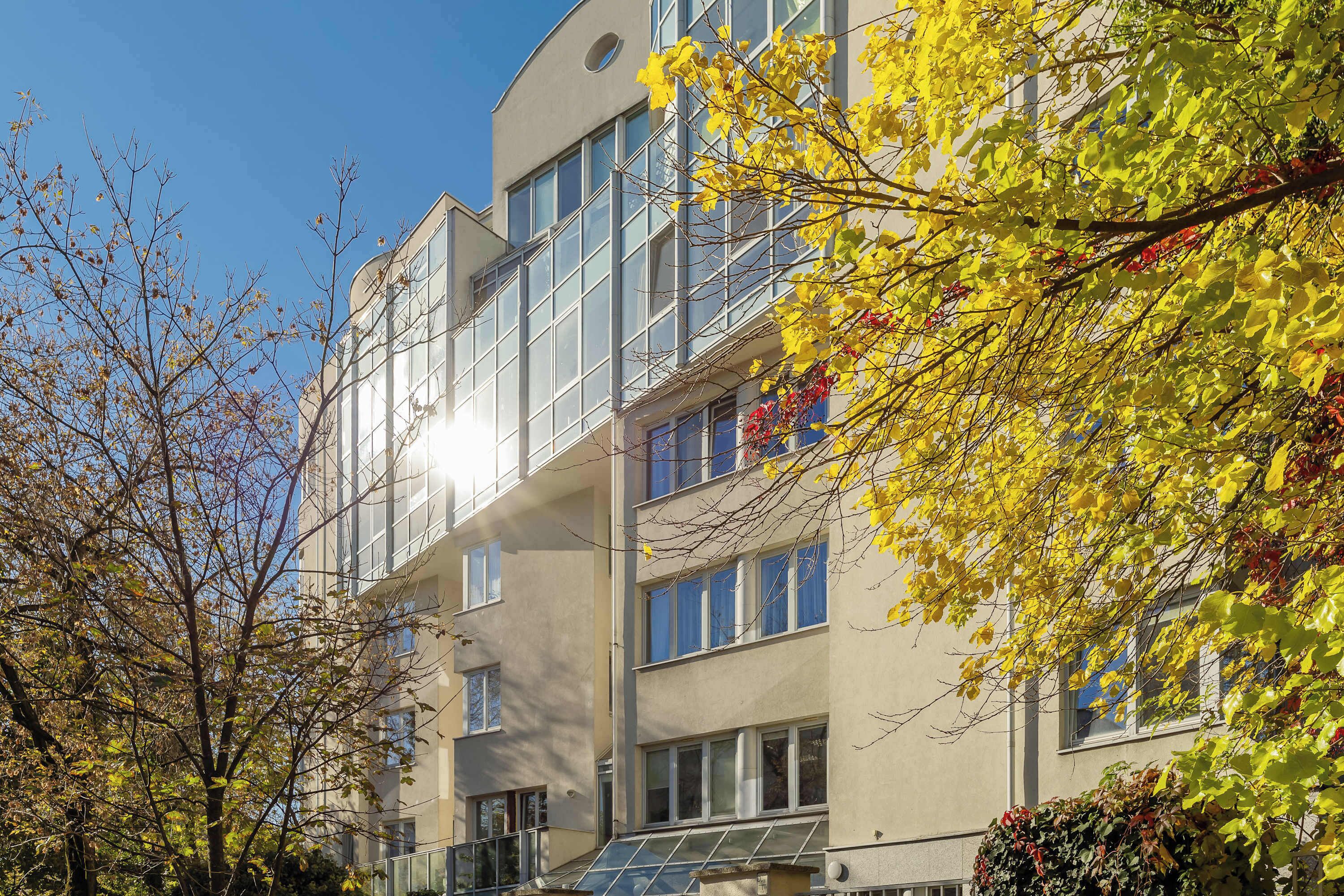 A multi-story building with large windows and balconies, surrounded by trees with yellow leaves.