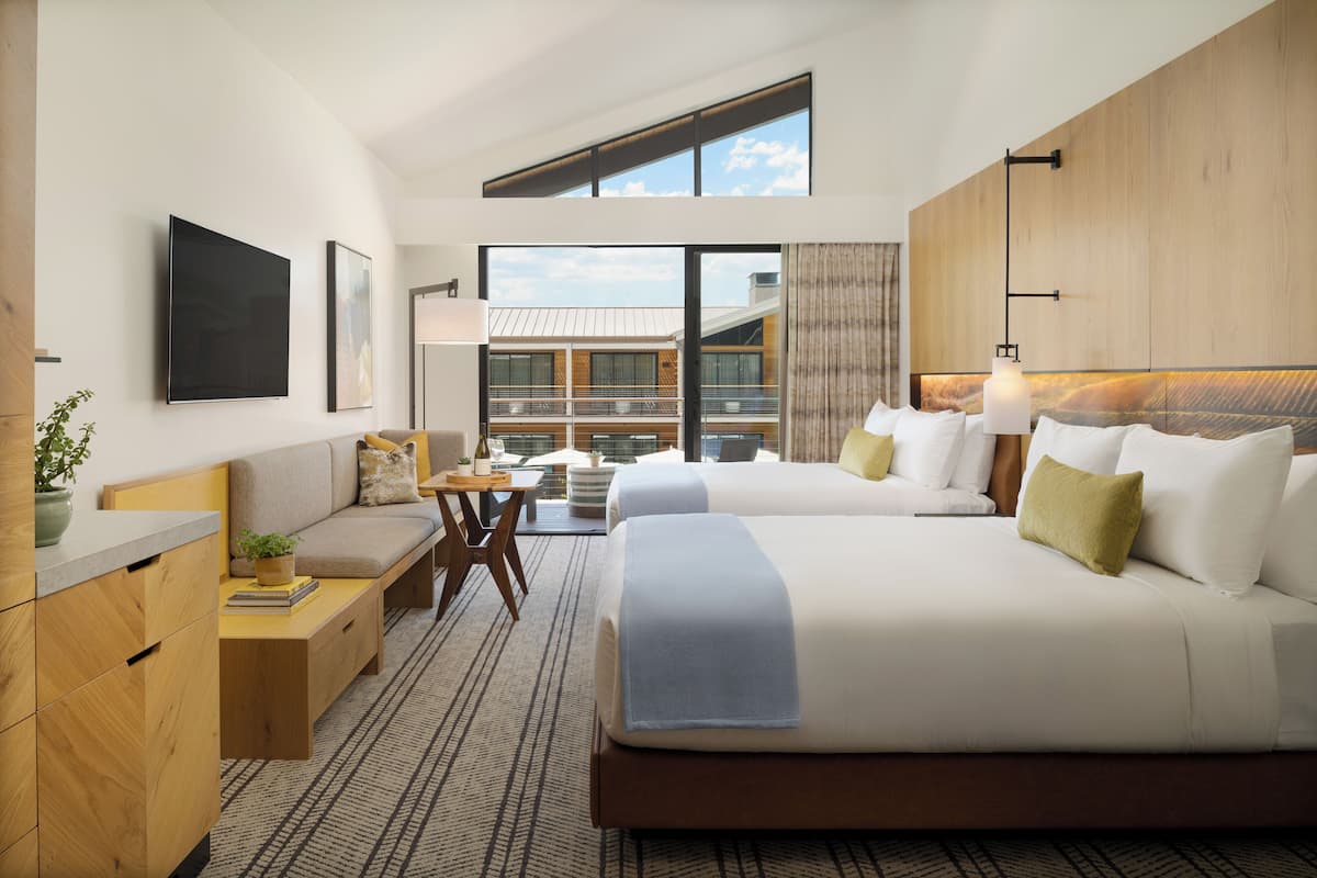 A modern hotel room with two beds, a desk, and a view of the building's exterior.