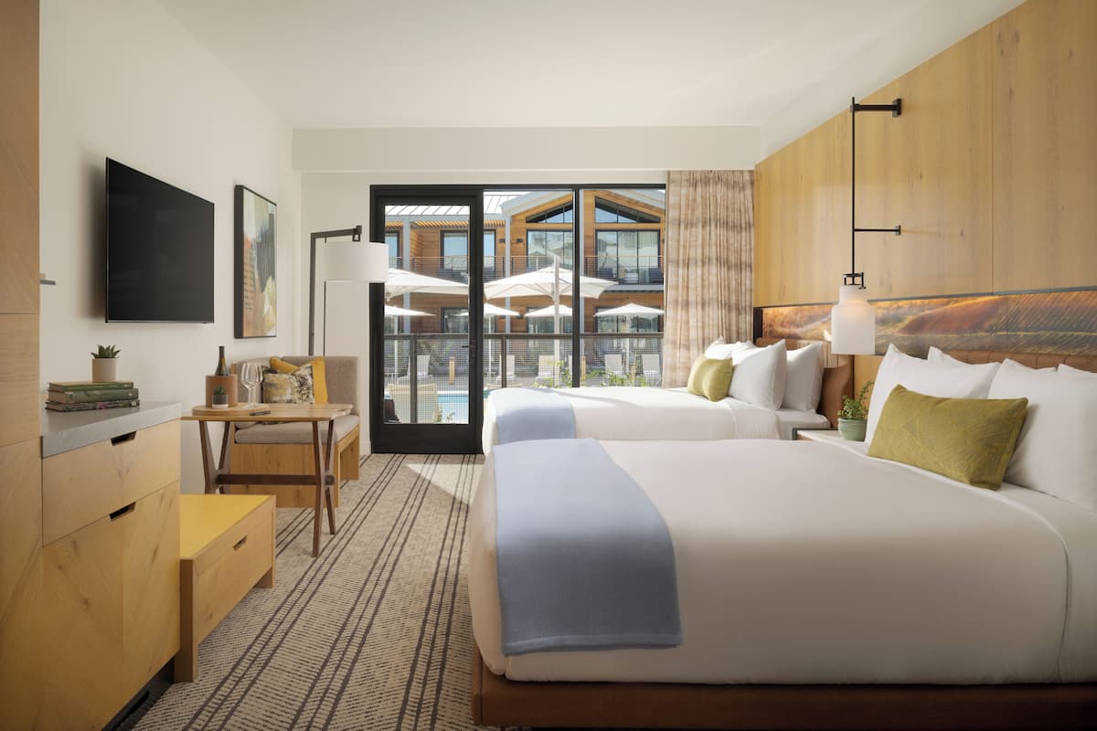 A hotel room with two beds, a desk, a TV, and a view of the outdoor area.