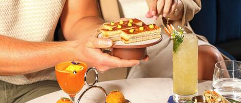 Une personne sert des plats à une table, notamment un sandwich composé de plusieurs couches, un cocktail agrémenté d'une garniture et un verre d'une boisson claire.