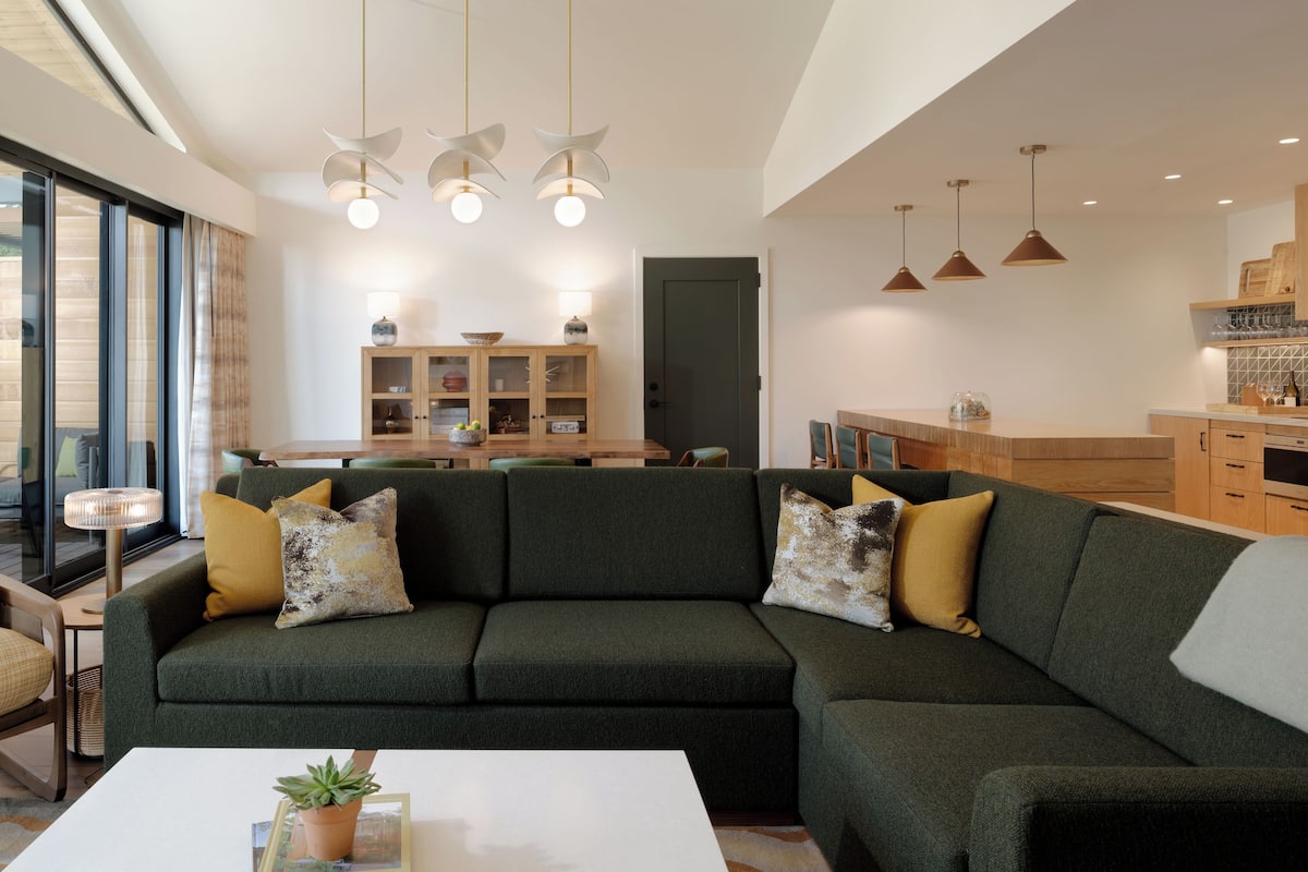 A modern living room with a green sofa, a white coffee table, and a dining area with wooden cabinets and pendant lights.