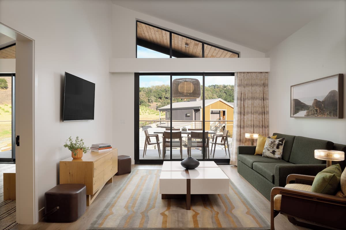 A modern living room with a large window, a flat-screen TV, a sofa, a coffee table, and a dining area with chairs and a table.