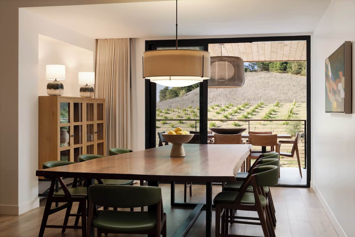 A dining area with a large wooden table, green chairs, and a bowl of lemons. There is a wooden cabinet with glass doors and a hanging light fixture.