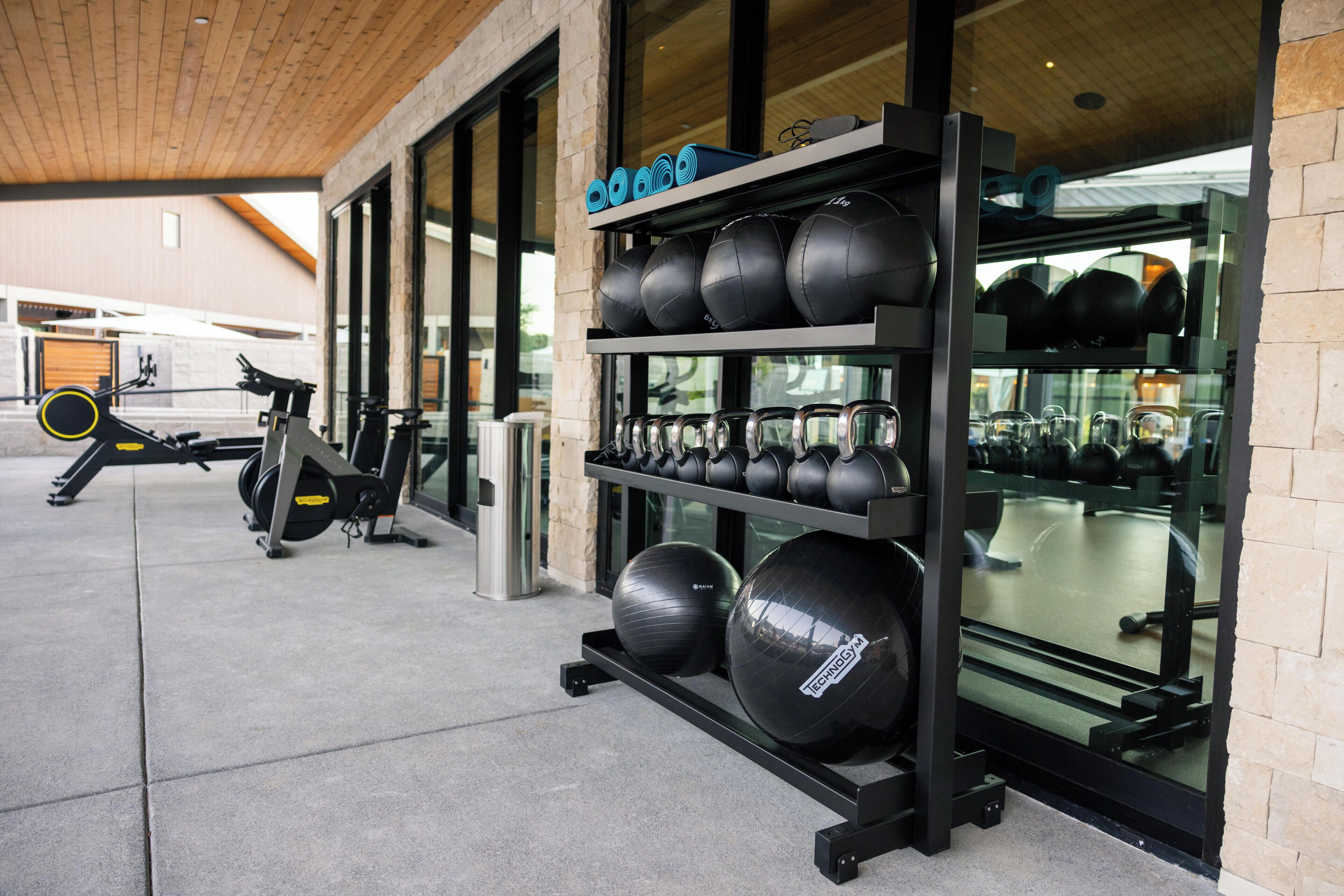 A gym with exercise equipment, a wall-mounted rack with kettlebells, and a window with a view of the street.