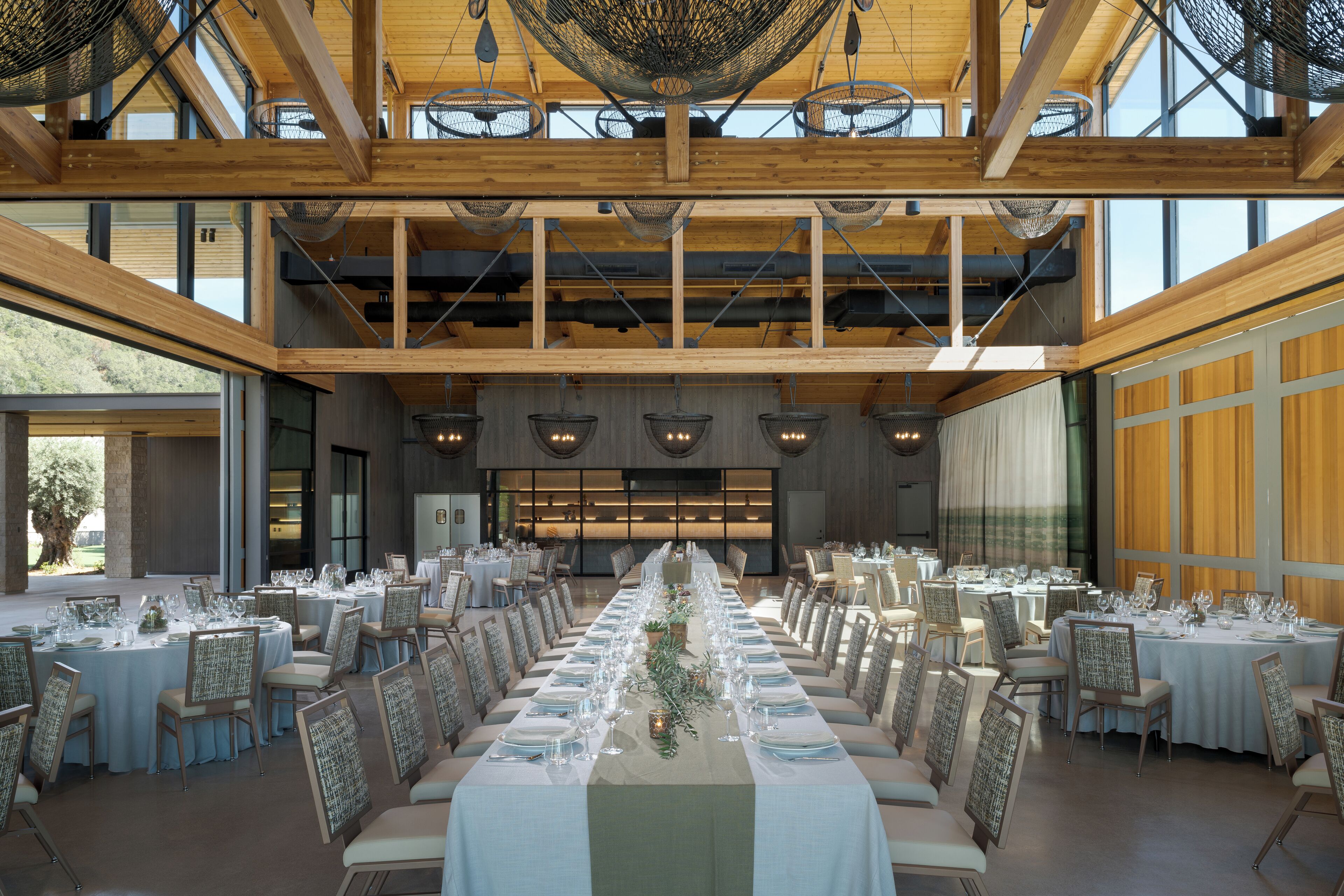 Uma sala de jantar ampla com longas mesas preparadas para um evento formal, tetos de madeira altos e grandes janelas que permitem entrada de luz natural.