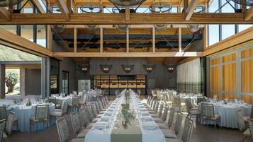 Uma sala de jantar ampla com longas mesas preparadas para um evento formal, tetos de madeira altos e grandes janelas que permitem entrada de luz natural.