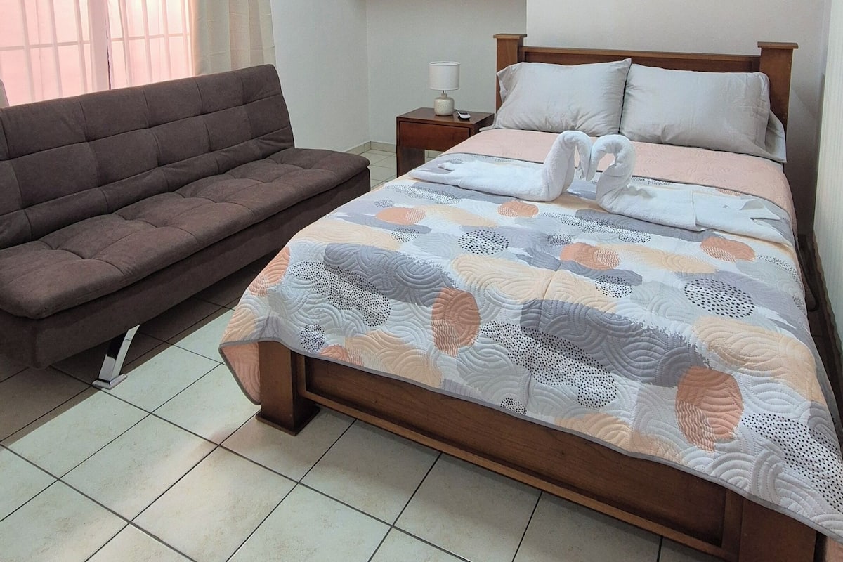 A hotel room with a bed, a sofa, a nightstand, and a wall-mounted air conditioner.
