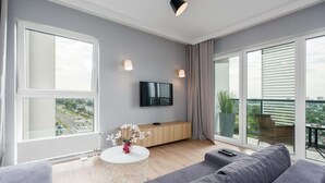 A modern living room with a grey sofa, a round coffee table with a vase of flowers, a flat-screen TV mounted on the wall, and large windows offering a city view.