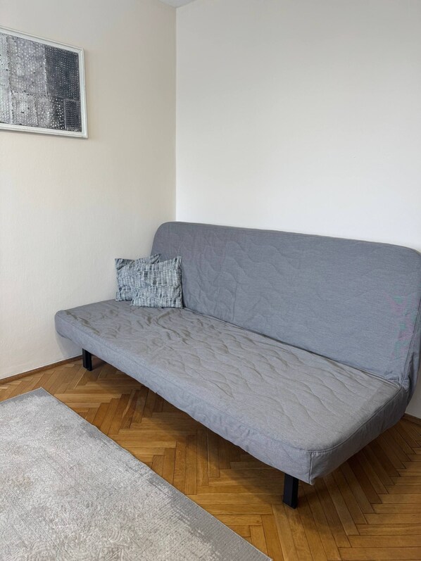 Un sofá gris con un cojín estampado, una alfombra gris y un suelo de madera con patrón de espiga.