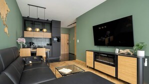 A modern living room with a black leather sofa, a dining area with a wooden table and chairs, a kitchen with dark cabinets and a black countertop, and a mounted flat-screen TV.
