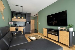 A modern living room with a black leather sofa, a dining area with a wooden table and chairs, a kitchen with dark cabinets and a black countertop, and a mounted flat-screen TV.