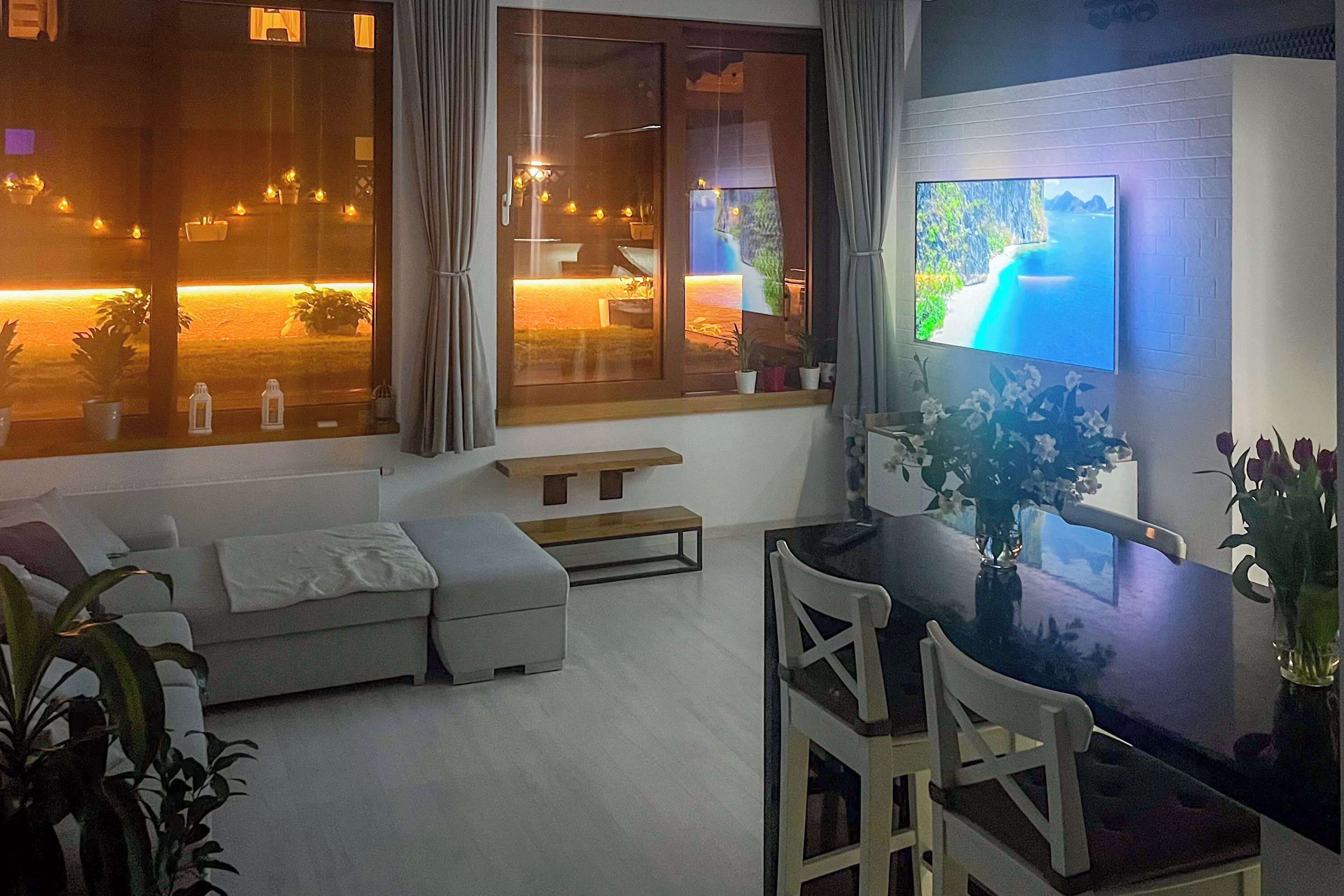 Uma sala de estar moderna com sofá, mesa de jantar e cadeiras, televisão fixada na parede e janelas amplas com cortinas.