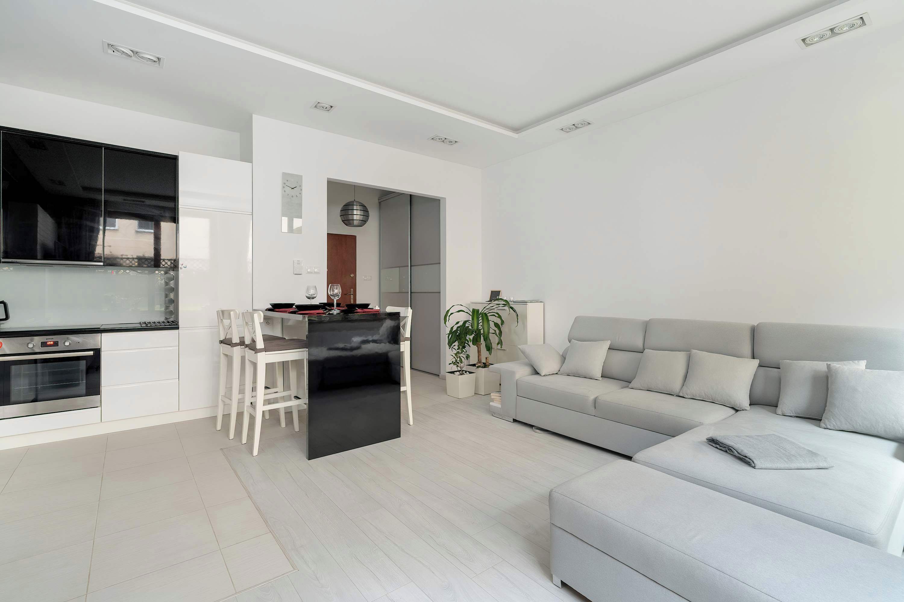 A modern living room with a grey sofa, a dining area with a black table, and a kitchen with white cabinets.