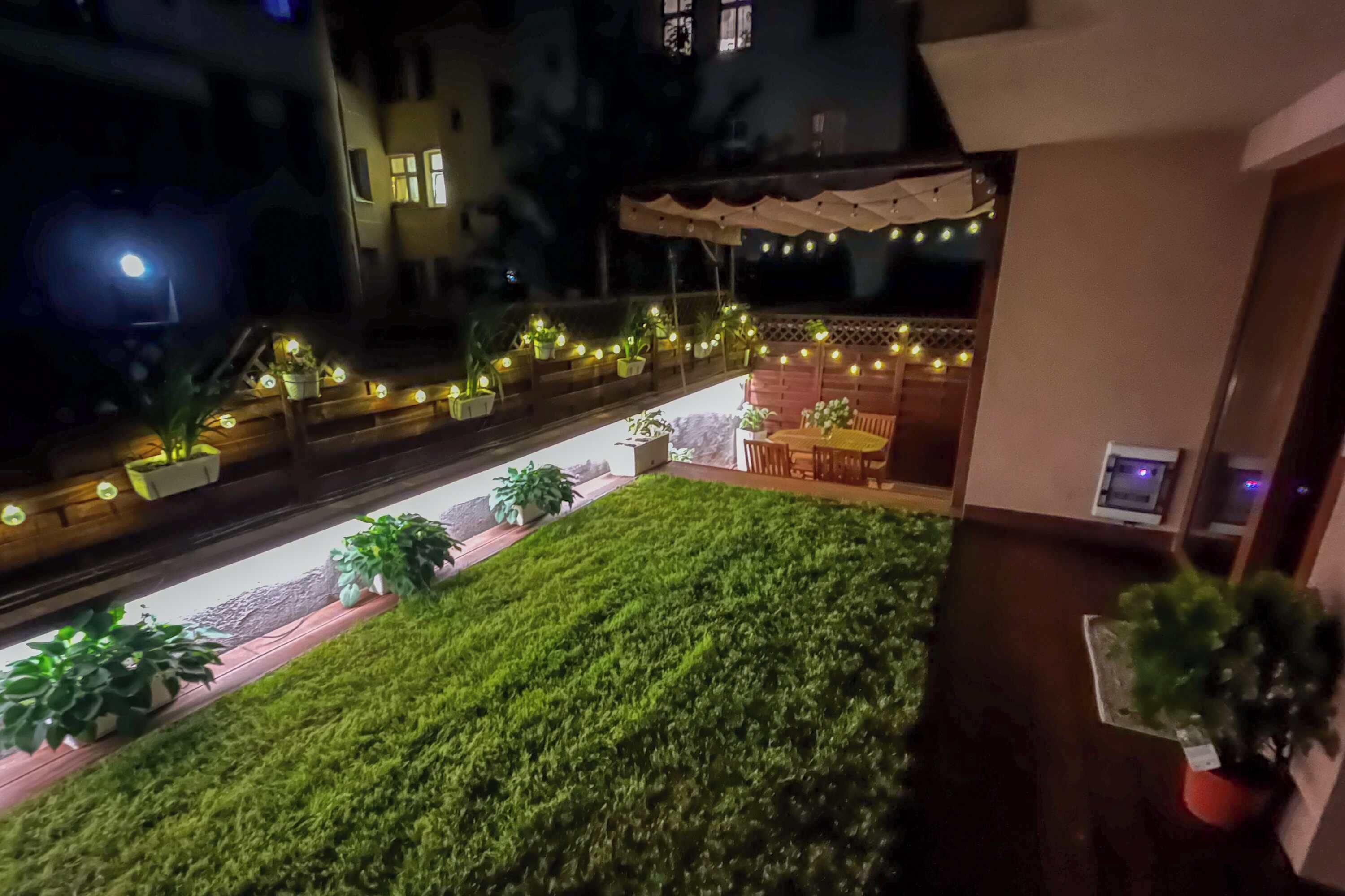Een terras op het dak met een verzorgd gazon, plantenpotten en lichtslingers.