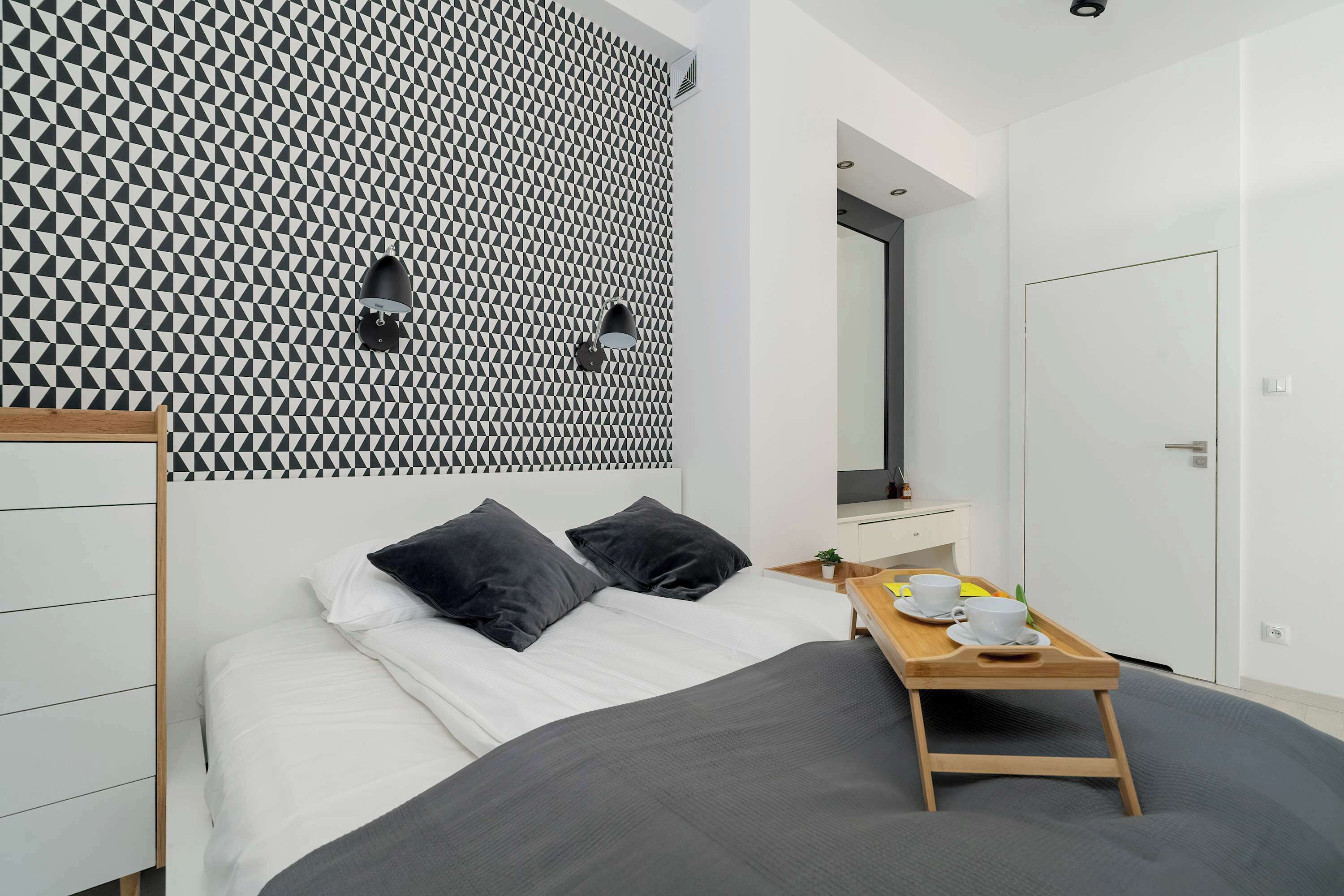 A modern bedroom with a geometric patterned wall, a bed with dark bedding, a bedside table with a tray, and a mirror above the bed.