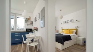 A modern bedroom with a bed, bedside tables, and a nightstand. There is a window with blinds, a white desk with a chair, and a small kitchen area with a microwave and a plant.