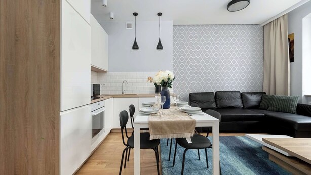 A modern kitchen with white cabinets, a dining table with chairs, a black leather sofa, and a patterned wall.