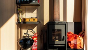 Una habitación acogedora con una estufa de leña, estanterías con libros y adornos, y una mesa con un objeto blanco y cables negros.