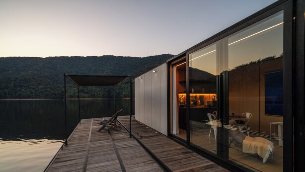 Una moderna casa flotante con terraza de madera, paredes de vidrio y vistas a un lago y colinas.