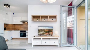 A modern living room with a sofa, a TV on a white cabinet, a sliding glass door, and a kitchen in the background.