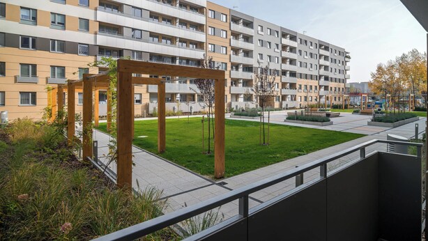 A multi-story residential building with balconies, a grassy courtyard, and a wooden pergola structure.