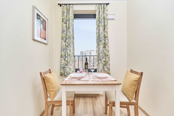A dining area with a table set for two, two chairs, a window with curtains, and a framed picture on the wall.