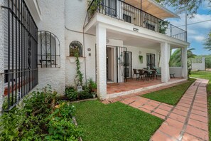 Un bâtiment blanc doté d’un balcon, une terrasse avec une table et des chaises, et un jardin bien entretenu.