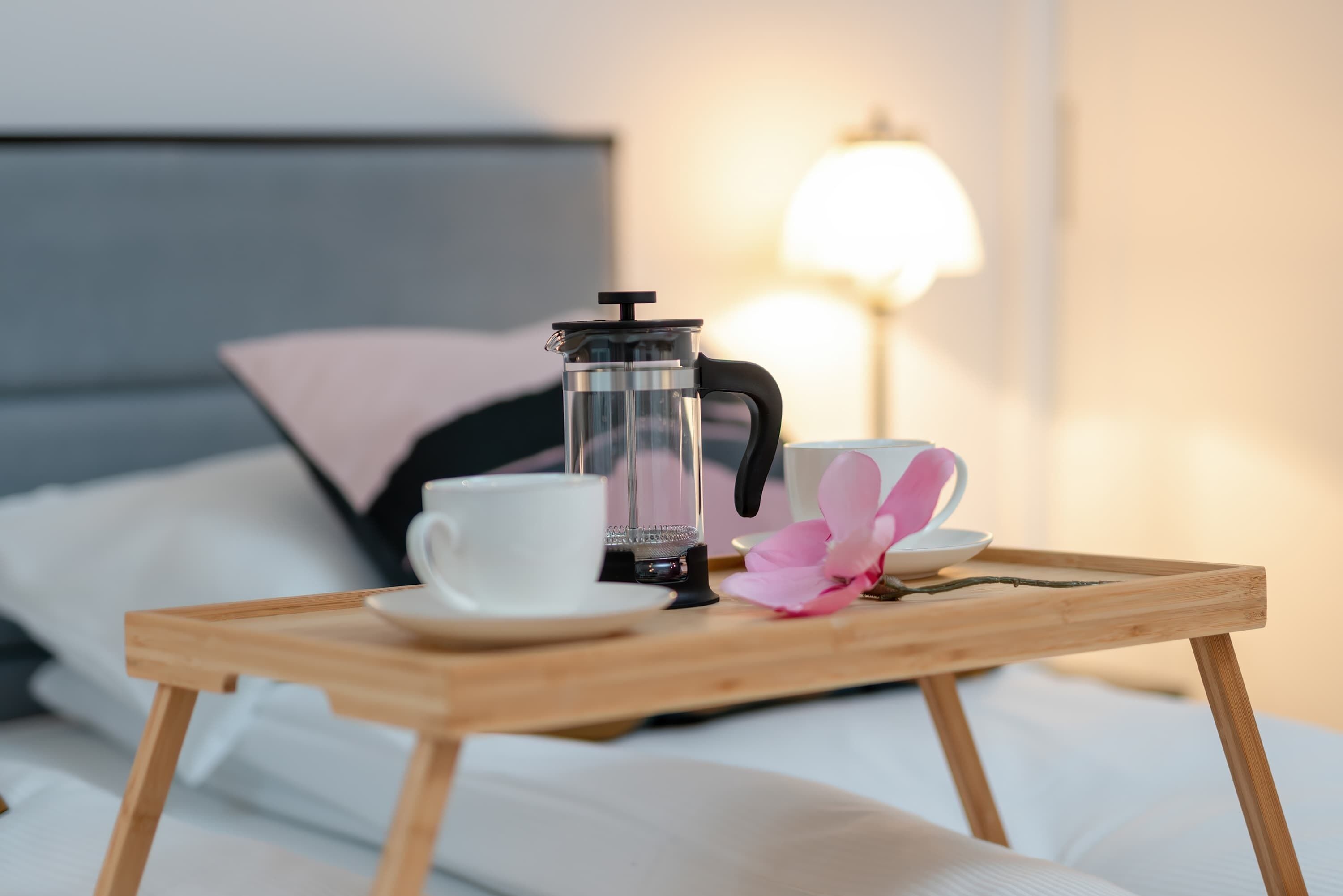 Puinen tarjotinalusta, jossa on ranskalainen kahvipannu, valkoinen kuppi ja vaaleanpunainen kukka. Taustalla on valkoisilla lakanoiden peitetty sänky ja lamppu.