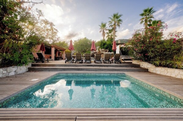 A swimming pool with lounge chairs and umbrellas, surrounded by greenery and palm trees.