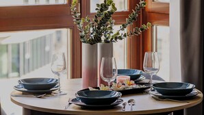 Mesa de comedor dispuesta con platos, vasos y un florero con plantas, ubicada junto a una ventana con cortinas.