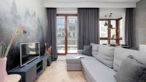 Una sala de estar moderna con un sofá gris, un televisor de pantalla plana sobre un soporte negro, piso con diseño de espiga y un gran ventanal con cortinas grises.