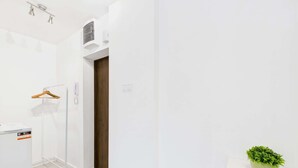 A hallway with a white wall, a wooden door, a hanging coat rack, a small potted plant, and a white cabinet with a red stripe.