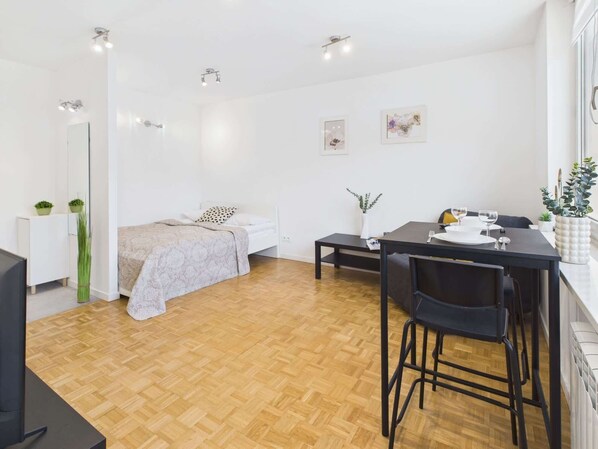 Een moderne, goed verlichte kamer met een bed, een eettafel met stoelen en een klein wit kastje.