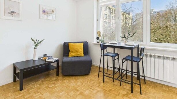 A modern living room with a sofa, a small table, and a bar stool. There are two windows with a view of buildings and trees.