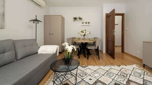 Una sala de estar con un sofá, una mesa de centro de vidrio, una mesa de comedor con sillas, una lámpara y una puerta que da a otra habitación.