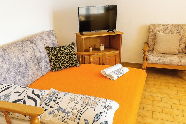 A living room with a sofa, a chair, a TV on a wooden stand, and a rolled-up towel on the floor.
