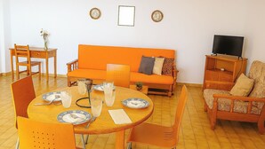 A living room with a round wooden dining table set for four, an orange sofa, and a television on a wooden stand. - Praia da Rocha 1 Bedroom Apartment With Balcony (Portimão)