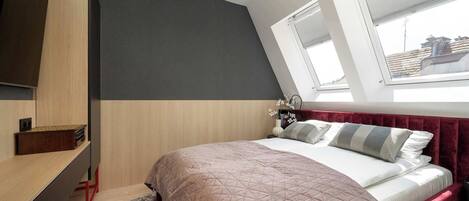 A bedroom with a bed, a desk, and a window with a view of buildings.