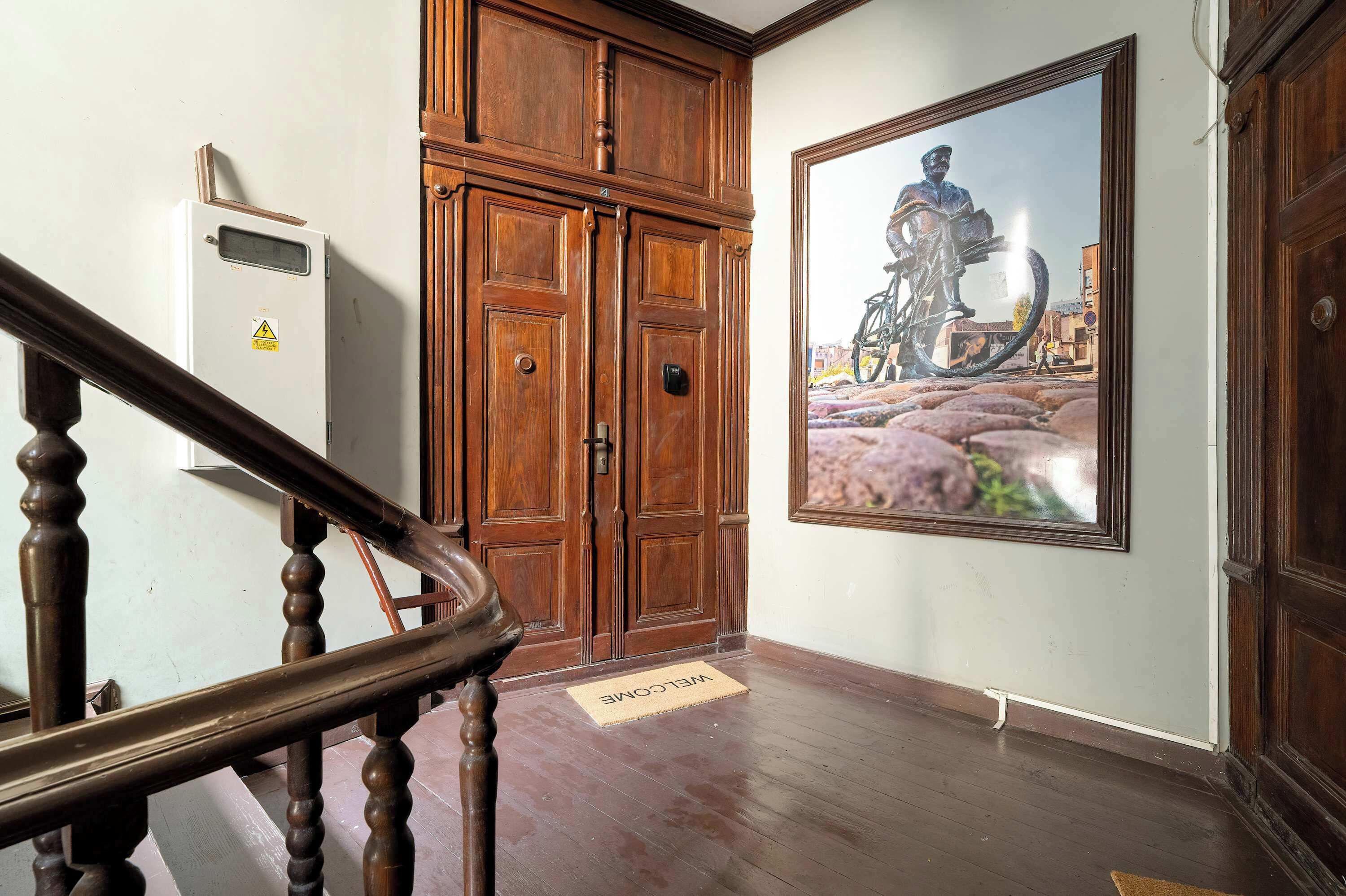 A wooden staircase with a welcome mat, a large wooden door, and a framed painting of a bicycle.