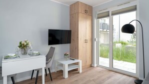 Een moderne kamer met een wit bureau, stoel en een aan de muur bevestigde televisie. Er is een houten kastje en een klein tafeltje met een bloemenvaas.
