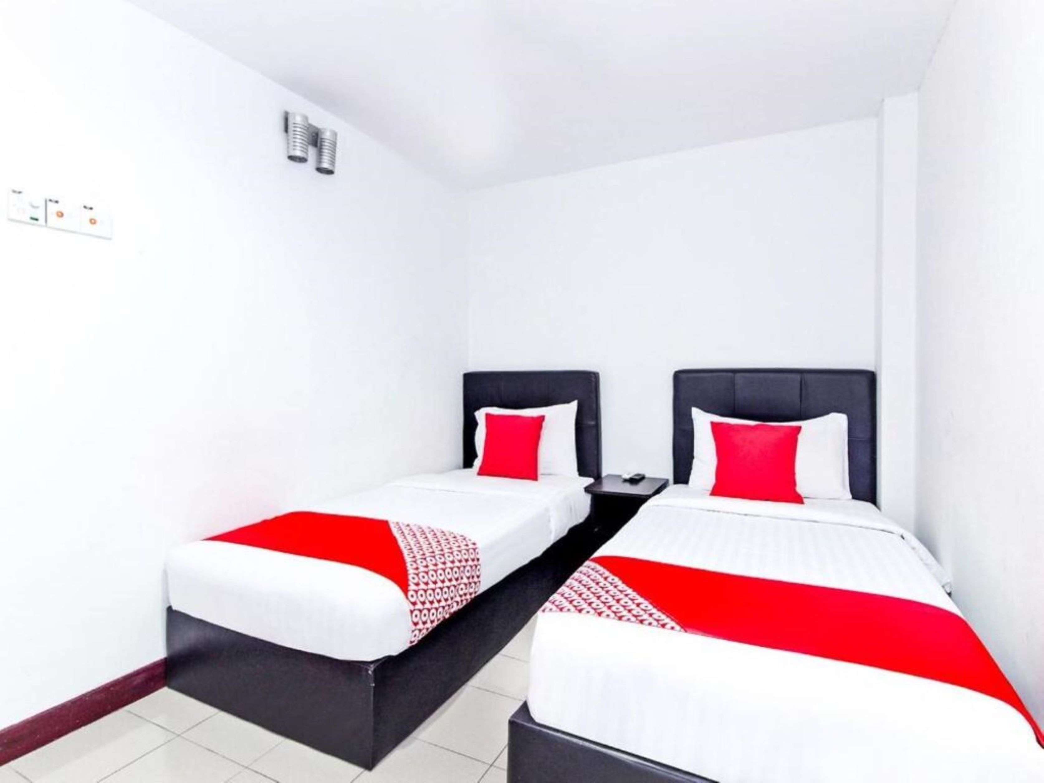 A hotel room with two single beds, each with a black headboard and red pillows. The room has white walls and a tiled floor.