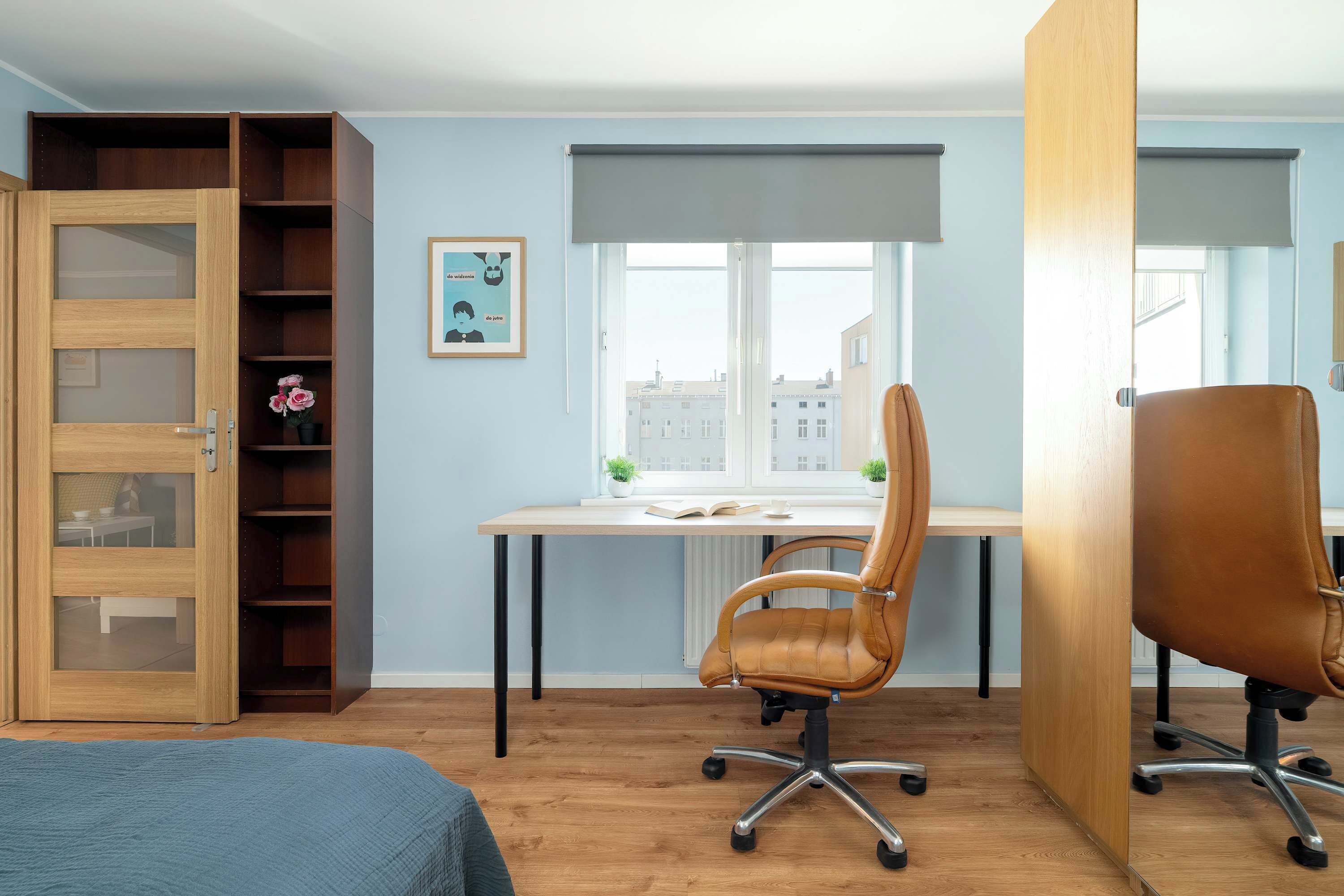 A bedroom with a wooden wardrobe, a desk with two chairs, a window with a view of buildings, and a bed with a blue cover.