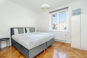 A bedroom with a bed, a small table, a wardrobe, and a window with a view of buildings. - Poznan | Rycerska 5 | Spacious Apartment | Free Parking (Poznań)