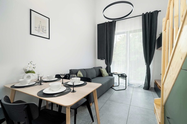 A modern studio with a functional kitchenette, an elegant table with four chairs, stylish lighting, and a large window letting in plenty of natural light.