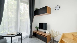 A modern living room with a wooden TV stand, a black-framed desk, a potted plant, and a clock on the wall.